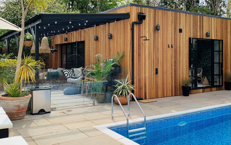 Wooden-clad backyard with glass walls, string lights, and potted plants; a blue-tiled pool with a metal ladder in the foreground and seating beyond.