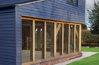 Blue two-story wooden building with a gabled roof, featuring a large glass-walled extension framed in light timber and a brick base on a green lawn.