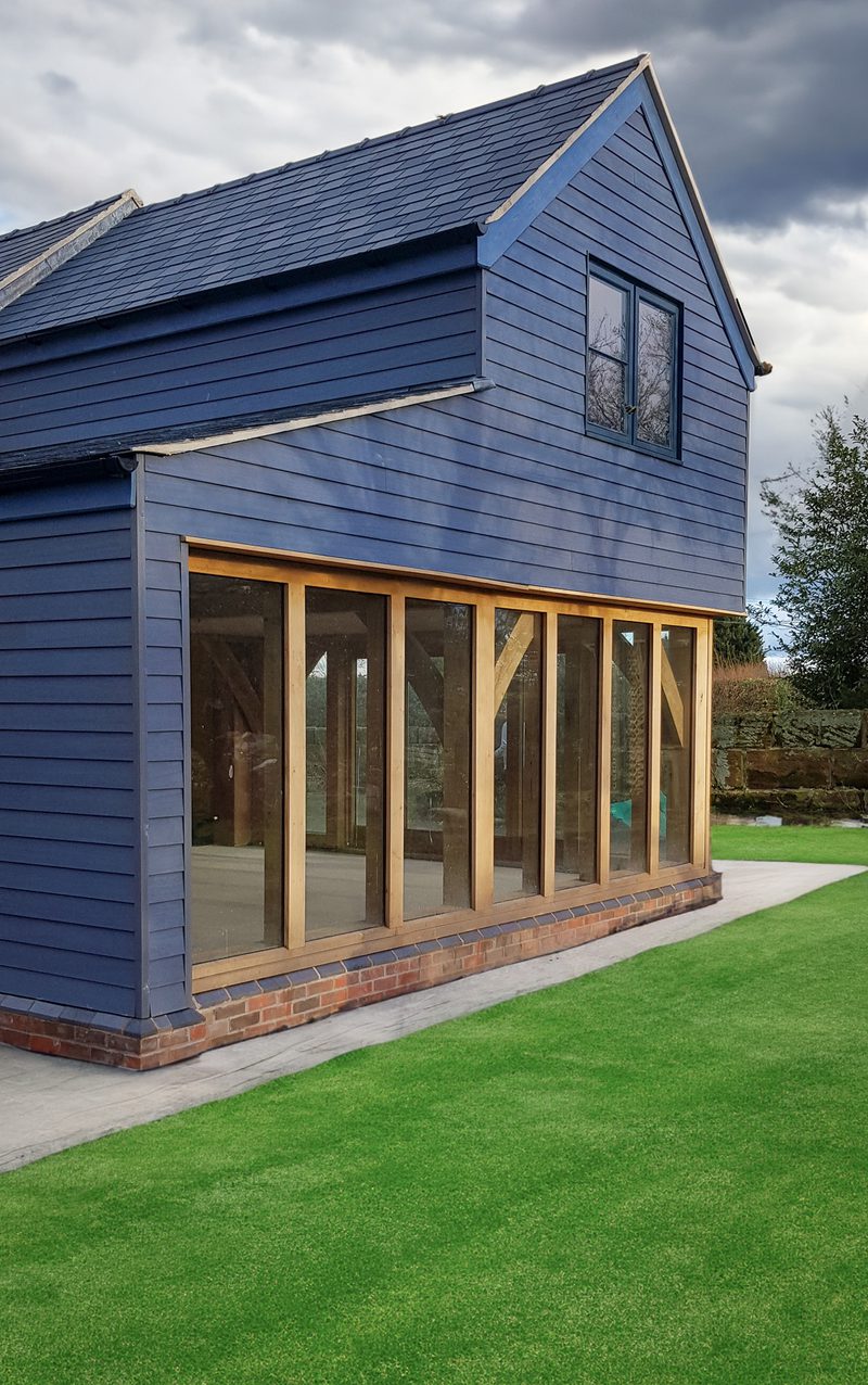 Blue two-story wooden building with a gabled roof, featuring a large glass-walled extension framed in light timber and a brick base on a green lawn.
