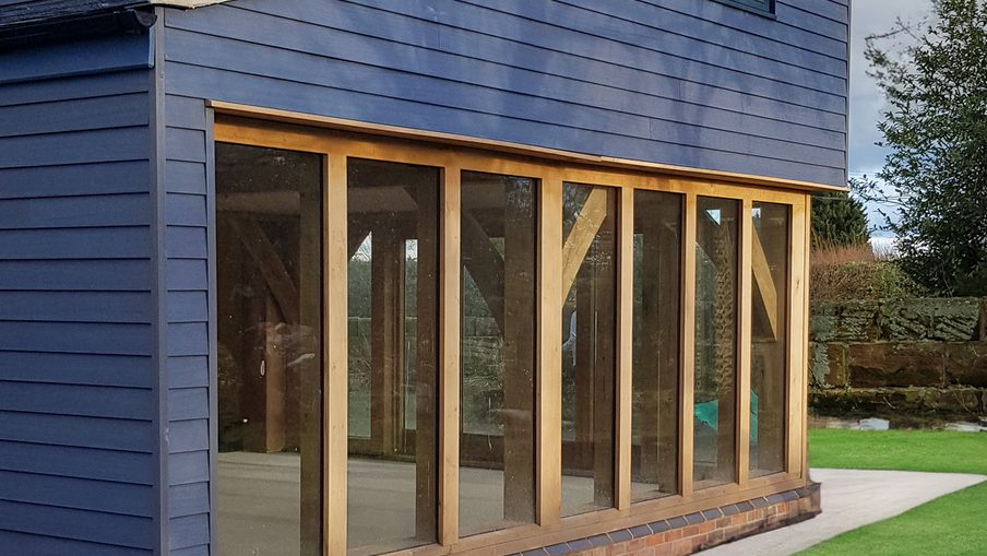 Blue two-story wooden building with a gabled roof, featuring a large glass-walled extension framed in light timber and a brick base on a green lawn.