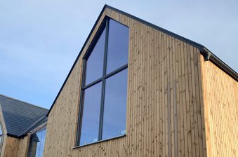 Modern wooden house with tall vertical planks and large glass panels; angular rooflines and a blue sky, sunlight along the right side.