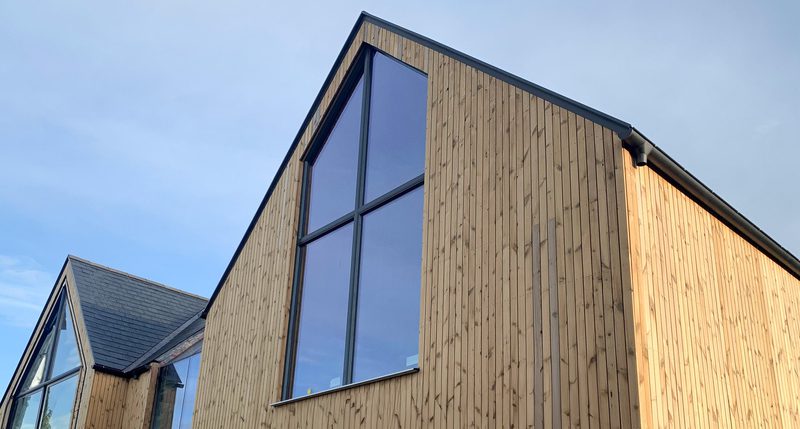 Modern wooden house with tall vertical planks and large glass panels; angular rooflines and a blue sky, sunlight along the right side.