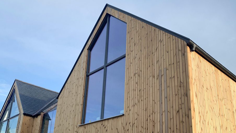 Modern wooden house with tall vertical planks and large glass panels; angular rooflines and a blue sky, sunlight along the right side.