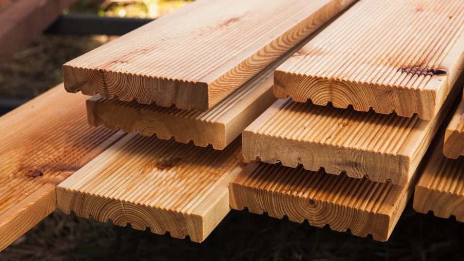 Stacks of pale pine boards with tongue-and-groove edges, outdoors on a grassy surface.