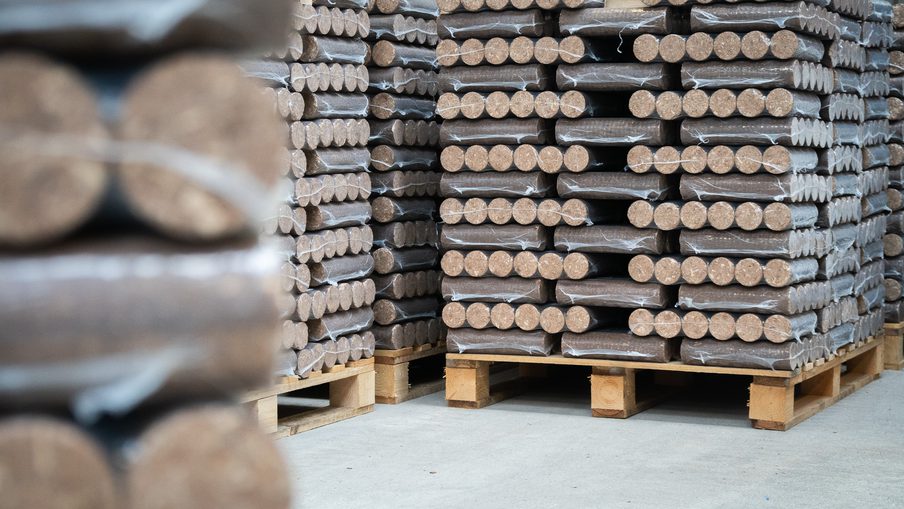 Stacks of round-ended wooden timbers wrapped in plastic, stacked on wooden pallets in a warehouse.