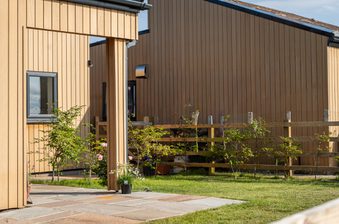 Two-toned wooden building with vertical tan siding, a small window, and a covered entry. Potted plants line a stone-paved patio beside a wooden fence and green lawn.