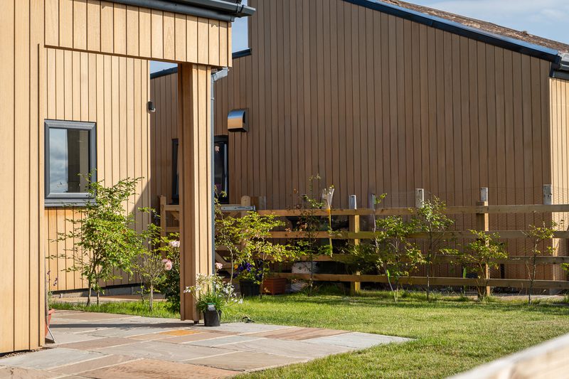 Two-toned wooden building with vertical tan siding, a small window, and a covered entry. Potted plants line a stone-paved patio beside a wooden fence and green lawn.