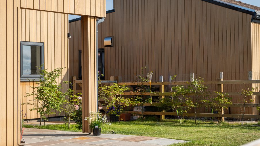 Two-toned wooden building with vertical tan siding, a small window, and a covered entry. Potted plants line a stone-paved patio beside a wooden fence and green lawn.