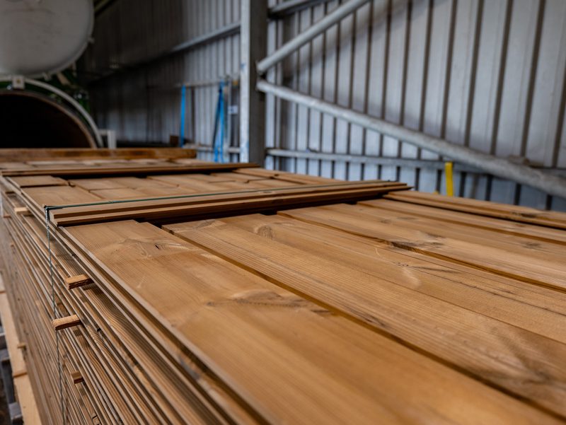 Stacks of wooden planks in a metal-walled warehouse, with industrial equipment and blue supports visible in the background.