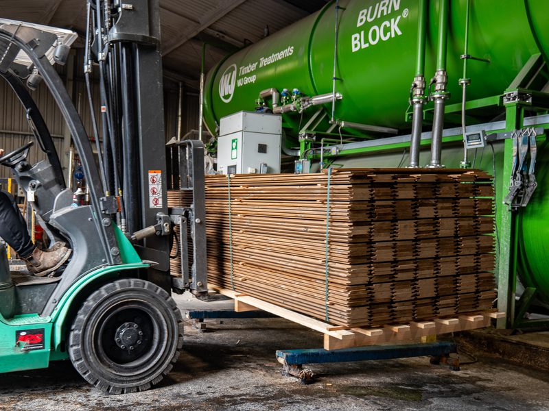 Forklift at a green industrial plant moves a pallet of stacked cardboard sheets beside a large green cylinder labeled Burn Block.