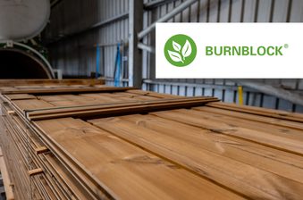 Stacked wooden planks in an industrial lumber warehouse; a white Burnblock logo sits in the upper-right corner.