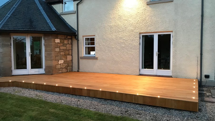 Elevated wooden deck along the back of a beige stucco house, with glass doors and a small window, a stone wall on the left, gravel border, and a lawn in front; edge lighting.