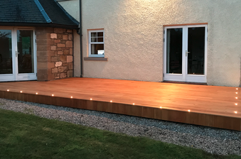 Wooden deck along the back of a beige house with glass doors and a small window; warm lights line the edge over a gravel bed and lawn.
