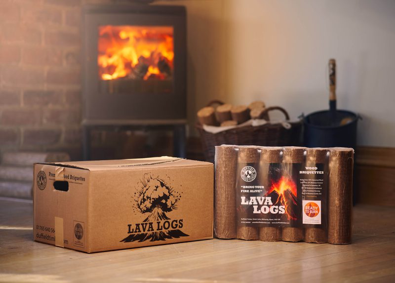 Two Lava Logs boxes on a wooden floor in front of a lit fireplace; a cardboard box with a volcano graphic and a six-pack of log bundles.