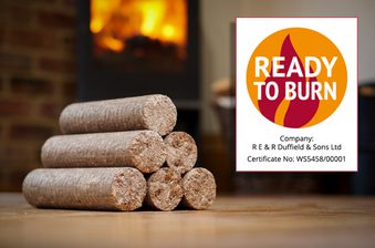 Stack of cylindrical wood logs on a wooden floor, with a lit fireplace in the background and a Ready to Burn certification sign on the right.