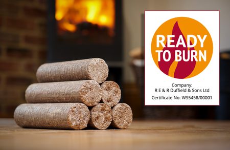 Stack of cylindrical wood logs on a wooden floor, with a lit fireplace in the background and a Ready to Burn certification sign on the right.