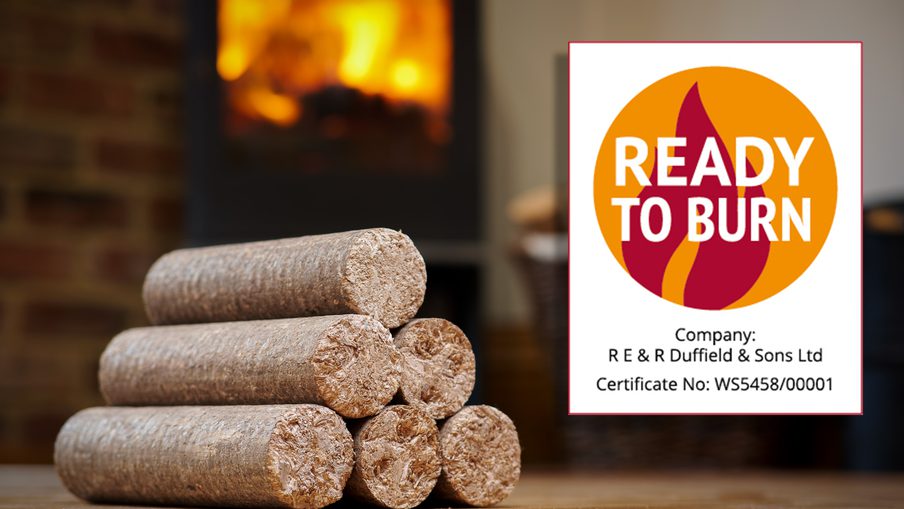 Stack of cylindrical wood logs on a wooden floor, with a lit fireplace in the background and a Ready to Burn certification sign on the right.