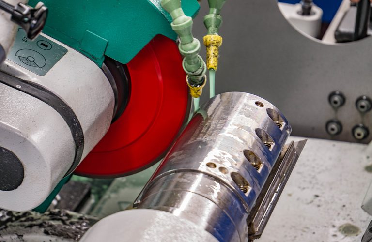 Close-up of a metal lathe with a polished cylindrical workpiece held in a chuck, red guard wheel, green housing, and yellow coolant hoses above.