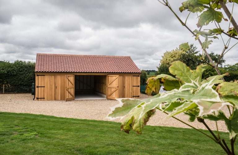 A wooden shed with a red-tiled roof sits on a gravel driveway, its double doors open. A green lawn and hedge frame the scene under a cloudy sky.