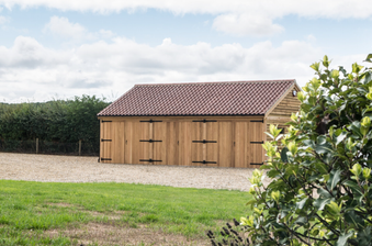 Wooden garden shed with a red-tiled roof sits on a gravel driveway, flanked by hedges, green lawn and a leafy shrub frame the scene under cloudy sky