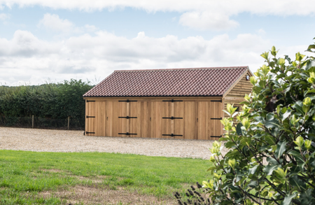 Wooden garden shed with a red-tiled roof sits on a gravel driveway, flanked by hedges, green lawn and a leafy shrub frame the scene under cloudy sky