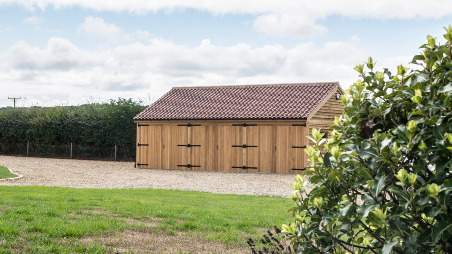 Wooden garden shed with a red-tiled roof sits on a gravel driveway, flanked by hedges, green lawn and a leafy shrub frame the scene under cloudy sky