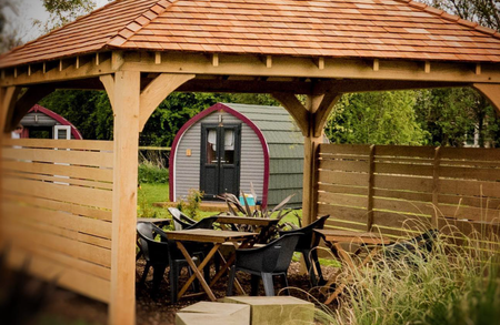 Wooden gazebo with a red-tiled roof shelters a table with black chairs; behind is a curved purple-trimmed shed near a wooden fence and greenery.