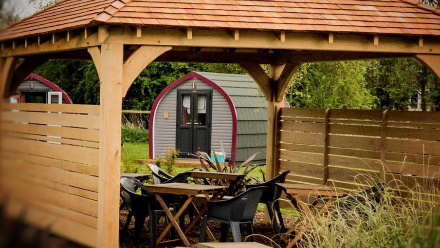 Wooden gazebo with a red-tiled roof shelters a table with black chairs; behind is a curved purple-trimmed shed near a wooden fence and greenery.