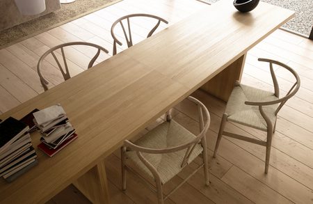 Bright minimalist dining area with a long light-wood table, pale woven chairs, a black bowl at the end, and magazines stacked on the side.