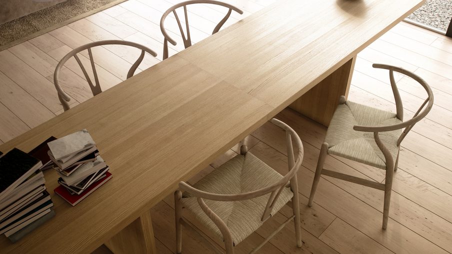 Bright minimalist dining area with a long light-wood table, pale woven chairs, a black bowl at the end, and magazines stacked on the side.