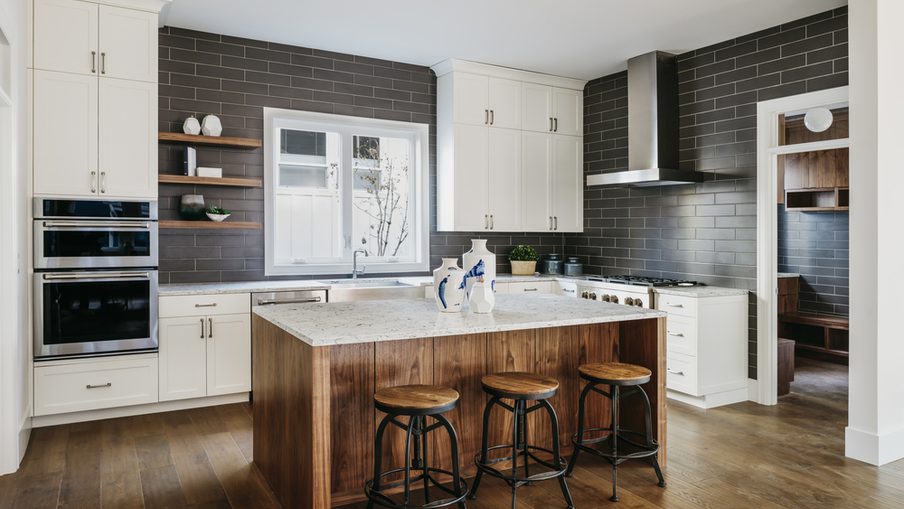 Bright modern kitchen with white shaker cabinets, dark subway-tile backsplash, marble-topped island with a wood base, three stools, stainless range hood, and double ovens.