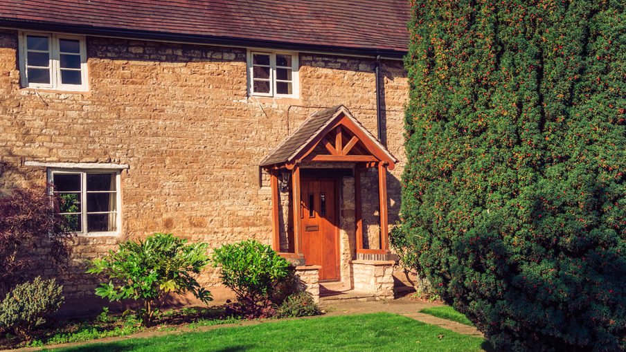 Beige stone cottage with a wooden front porch and door, a green lawn and shrubs in front, and a tall evergreen hedge on the right.