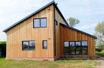 Modern wooden house with vertical siding, brick foundation, and large black-framed windows and doors; sits on a grassy yard under a blue sky.
