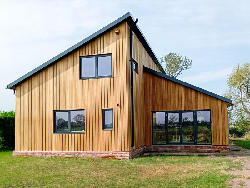 Modern wooden house with vertical siding, brick foundation, and large black-framed windows and doors; sits on a grassy yard under a blue sky.