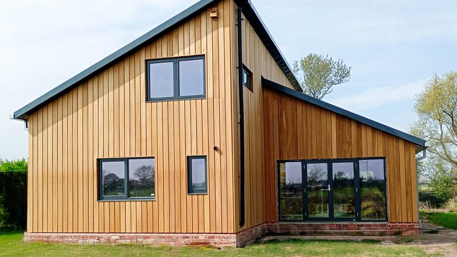 Modern wooden house with vertical siding, brick foundation, and large black-framed windows and doors; sits on a grassy yard under a blue sky.
