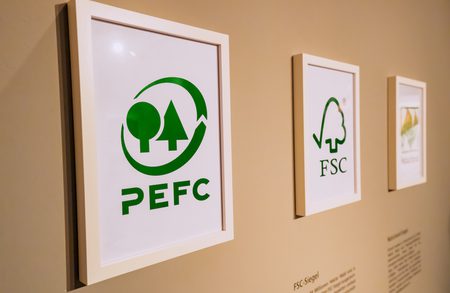 Three framed posters on a beige wall featuring forest-certification logos: left PEFC with tree icons, middle FSC logo, right another green emblem.