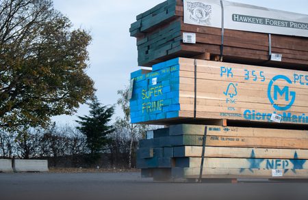 Stacks of lumber on pallets outdoors, blue-painted boards marked SUPER PRIME, with Hawkeye Forest Products banner and trees in the background.