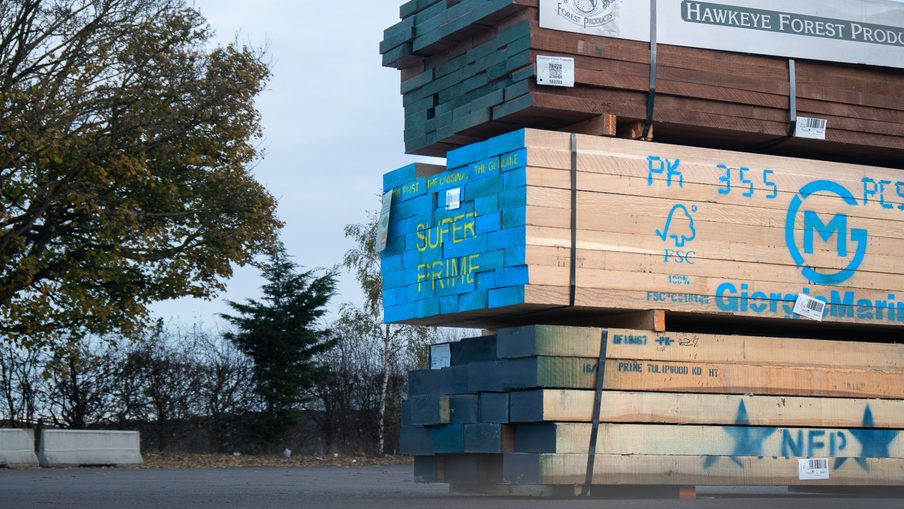 Stacks of lumber on pallets outdoors, blue-painted boards marked SUPER PRIME, with Hawkeye Forest Products banner and trees in the background.