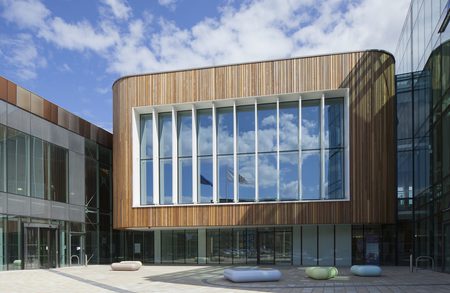 Modern building with curved wooden facade and a window with mullions; glass surrounds a tiled plaza with rounded pastel benches under blue sky.