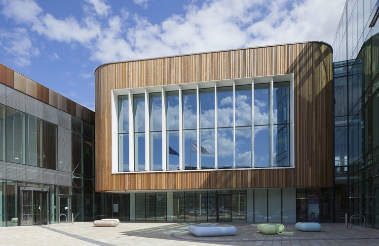 Modern building with curved wooden facade and a window with mullions; glass surrounds a tiled plaza with rounded pastel benches under blue sky.