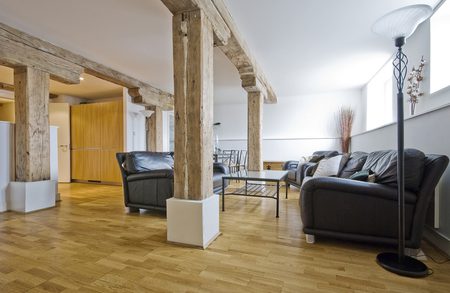 Loft-style living area with exposed wooden beams, black leather sofas, a glass coffee table, and a floor lamp; wooden floors, large windows, and a back kitchen.