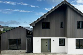 Modern two-story home with dark vertical siding atop a white base, a gray front door, several windows, under a blue sky with distant hills.
