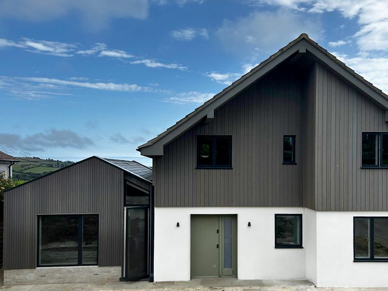 A modern house in Dorset, on the south coast of England, featuring Thermo-Ayous timber cladding in 'stone grey' with black-framed windows and a green front door.