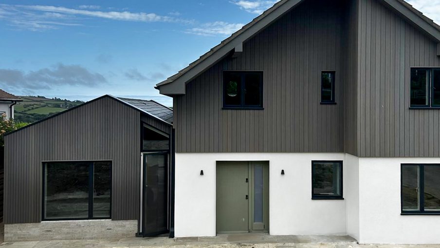 Modern two-story home with dark vertical siding atop a white base, a gray front door, several windows, under a blue sky with distant hills.