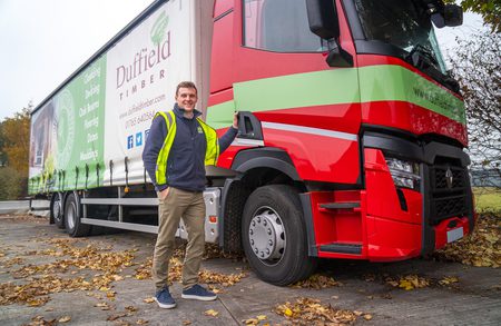 Person in a high-visibility jacket stands beside a large red and green Duffield Timber truck, autumn leaves on the ground.