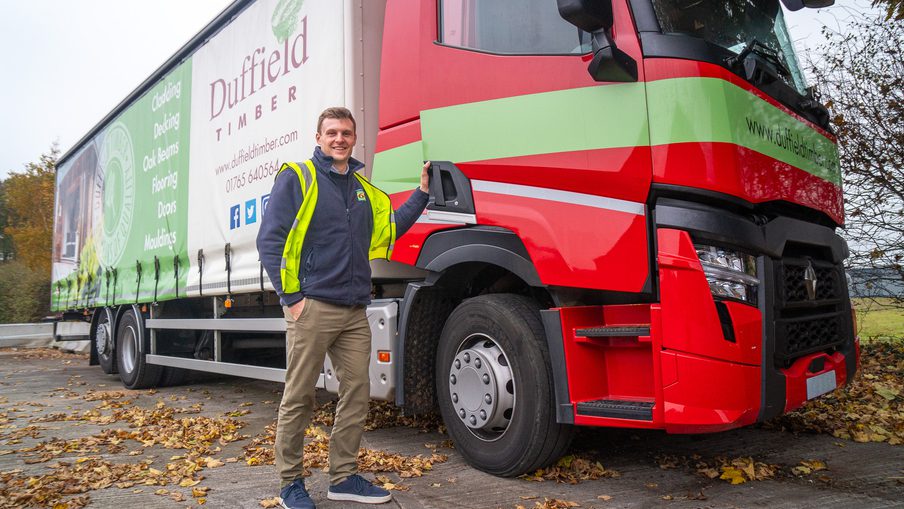 Person in a high-visibility jacket stands beside a large red and green Duffield Timber truck, autumn leaves on the ground.