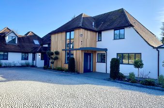 Modern white house with wood-clad entrance, dark roof, blue front door, large windows; cobblestone driveway and neatly trimmed shrubs under a clear blue sky.