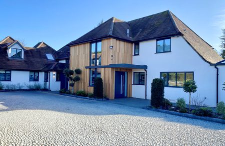 Modern white house with wood-clad entrance, dark roof, blue front door, large windows; cobblestone driveway and neatly trimmed shrubs under a clear blue sky.