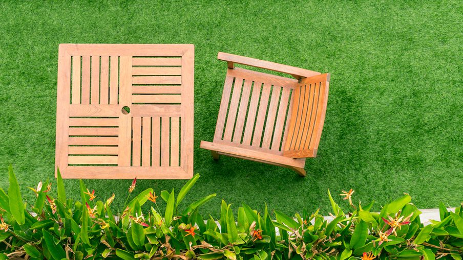 Overhead view of a wooden slatted chair and square table on a green lawn, with tropical plants along the bottom edge.