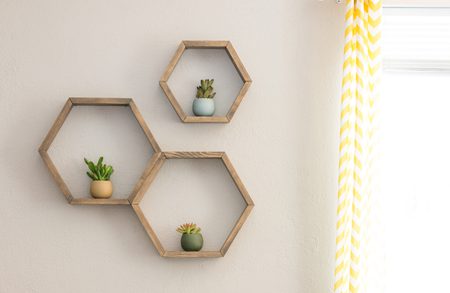 Three hexagonal wooden shelves on a beige wall, each holding a small succulent; bright window with a yellow-and-white striped curtain on the right.